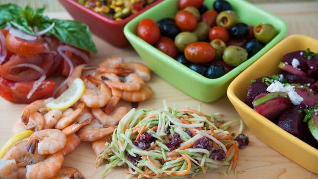 Shrimp, coleslaw and tomatoes in front of 3 containers holding olives, beets and corn